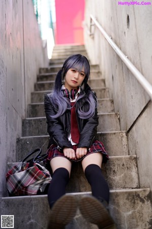 A woman in a plaid skirt and leather jacket leaning against a pole.